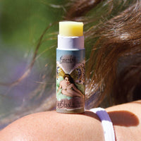 A tube of Barefoot Venus The Vanilla Effect Lip Balm, showcasing a natural lip balm design with a fairy illustration on the label, is uncapped and positioned on someone's shoulder. The moisturizing Shea Butter balm is highlighted under sunlight, with their hair visible in the background.