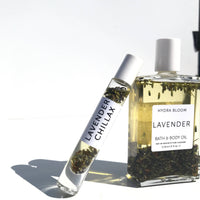 A clear roller bottle labeled "lavender chillax" beside a square glass bottle of "Hydra Bloom Beauty Luxe Lavender Bath and Body Oil" with lavender stems inside, against a white background with.
