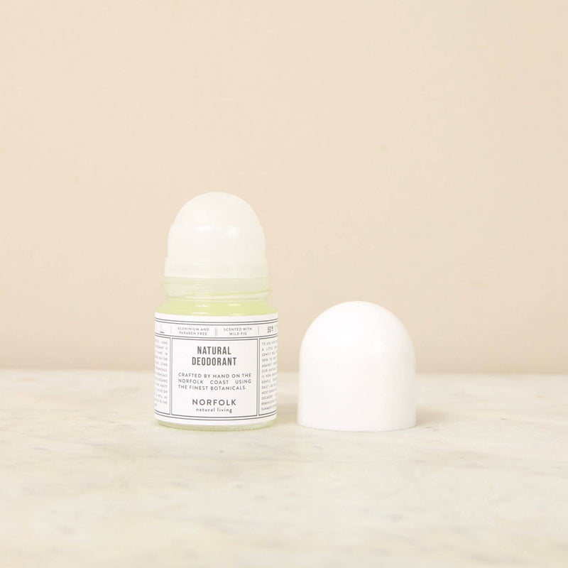 Against a plain beige background, the Norfolk Natural Living Fig Natural Deodorant for sensitive skin is displayed in a transparent bottle with a white label and cap. The label clearly reads "Norfolk Natural Living" and "Natural Deodorant," proudly highlighting its vegan-friendly origin and composition. The bottle sits beside its detached cap for emphasis.