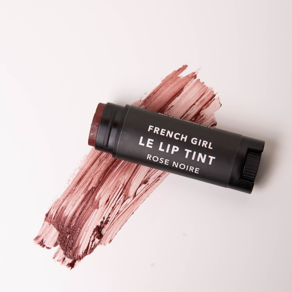 A FRENCH GIRL Tinted Lip Balm in Rose Noire rests on a light surface, with a swipe of its rich dark rose tint displayed underneath.