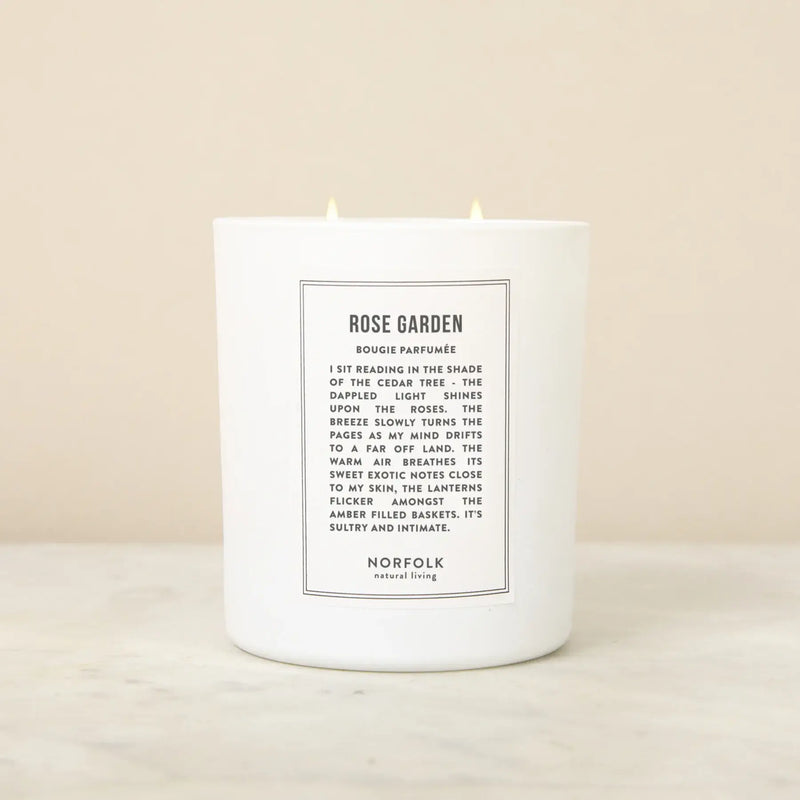 The Norfolk Natural Living Rose Garden Candle, 8oz, with two lit wicks sits on a light marble surface. The white botanical candle features a “Rose Garden” label and rests against a plain beige background.