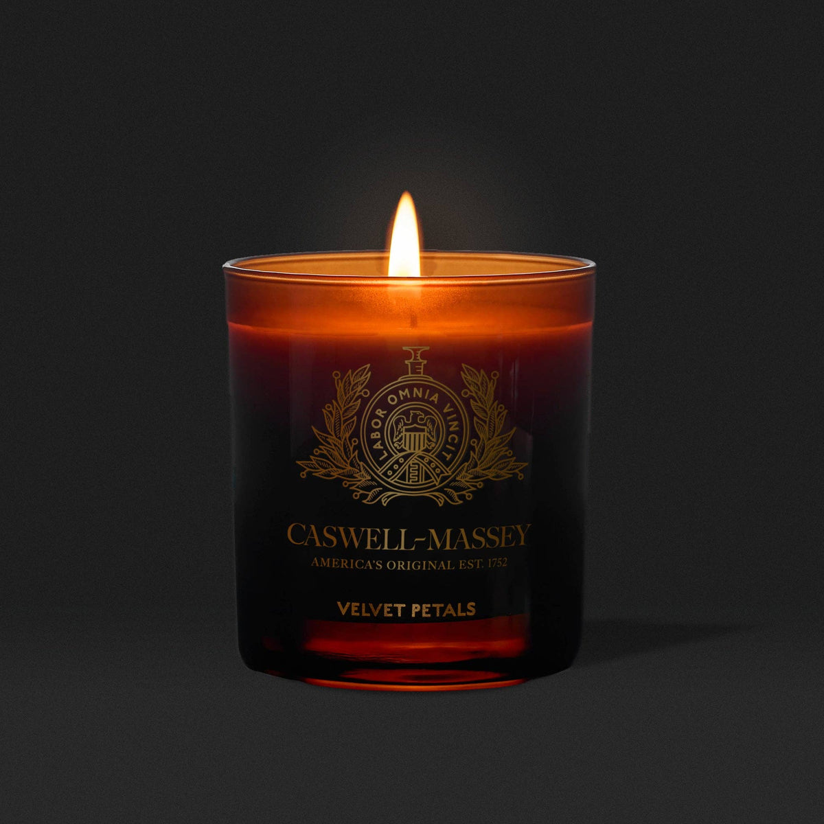 The Caswell-Massey Candle – Velvet Petals in a dark amber glass fills the air with a warm, floral scent, casting a cozy glow against a solid black background.