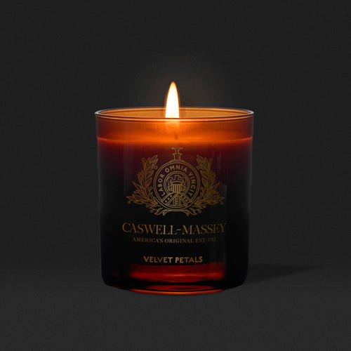 The Caswell-Massey Candle – Velvet Petals in a dark amber glass fills the air with a warm, floral scent, casting a cozy glow against a solid black background.