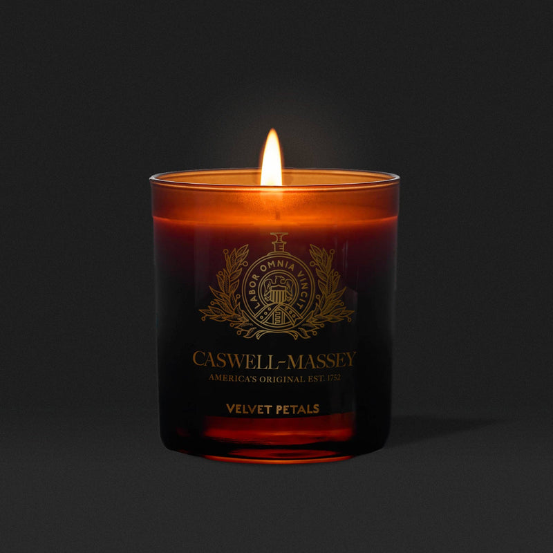 The Caswell-Massey Candle – Velvet Petals in a dark amber glass fills the air with a warm, floral scent, casting a cozy glow against a solid black background.