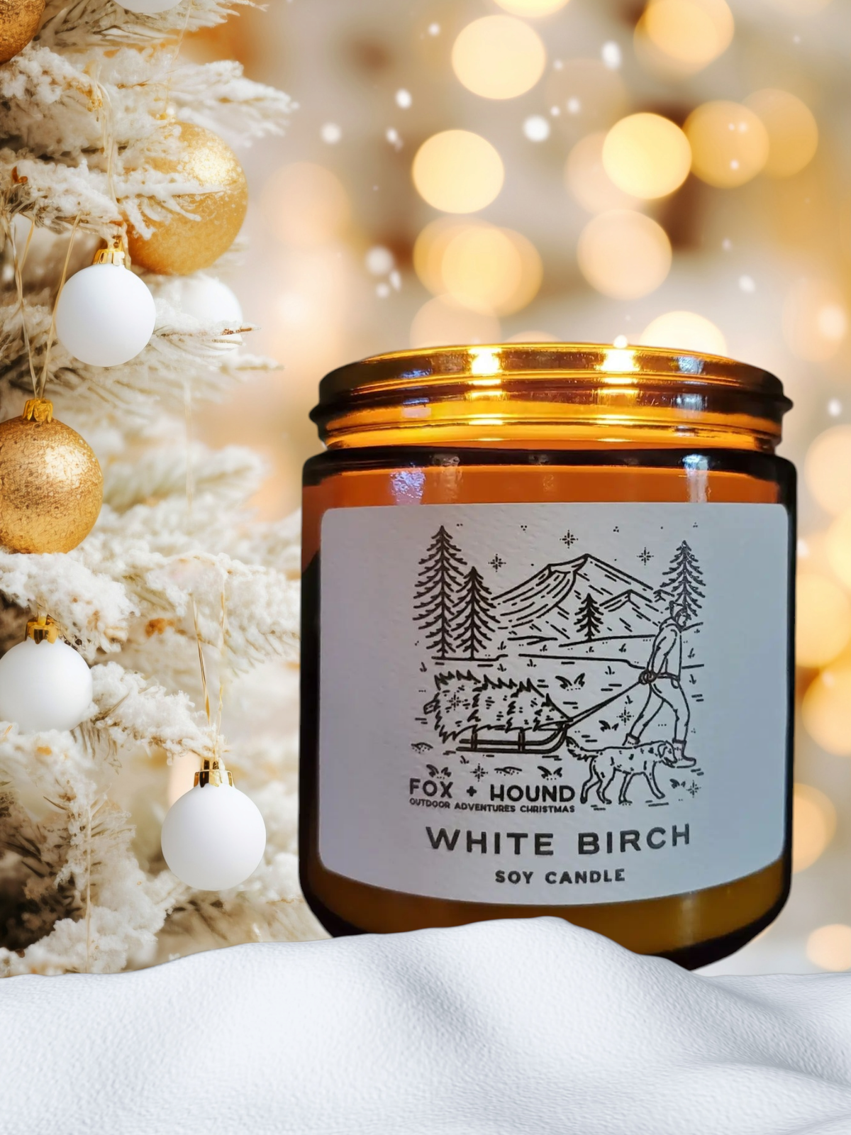 A Fox + Hound Odor Eliminator Soy Candle - White Birch in an amber jar rests beside a white Christmas tree with gold and white ornaments, as soft, warm lights glow gently in the background.