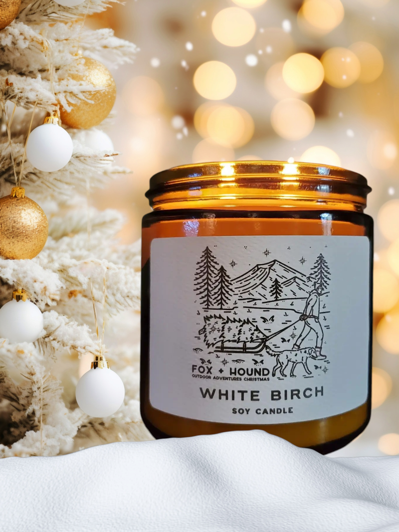 A Fox + Hound Odor Eliminator Soy Candle - White Birch in an amber jar rests beside a white Christmas tree with gold and white ornaments, as soft, warm lights glow gently in the background.