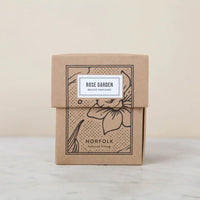 A brown box with floral illustrations and a white label displays the "Norfolk Natural Living Rose Garden Candle 8oz," featuring a botanical scent and shown on a light marble surface.