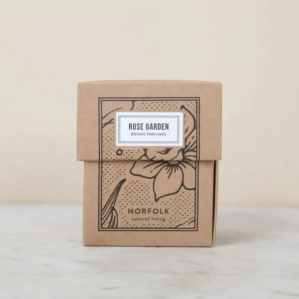 A brown box with floral illustrations and a white label displays the "Norfolk Natural Living Rose Garden Candle 8oz," featuring a botanical scent and shown on a light marble surface.