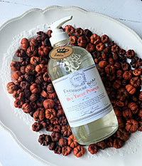 A bottle of "Z&Co. Hey There Pumpkin Farmhouse Fall Soap" is displayed on a white ornate platter filled with dried miniature pumpkins. The soap, featuring cocamidopropyl betaine, has a white pump and a decorative label with intricate designs and text. The rustic setting exudes an autumn vibe, perfect for pumpkin pie and creamy vanilla lovers.