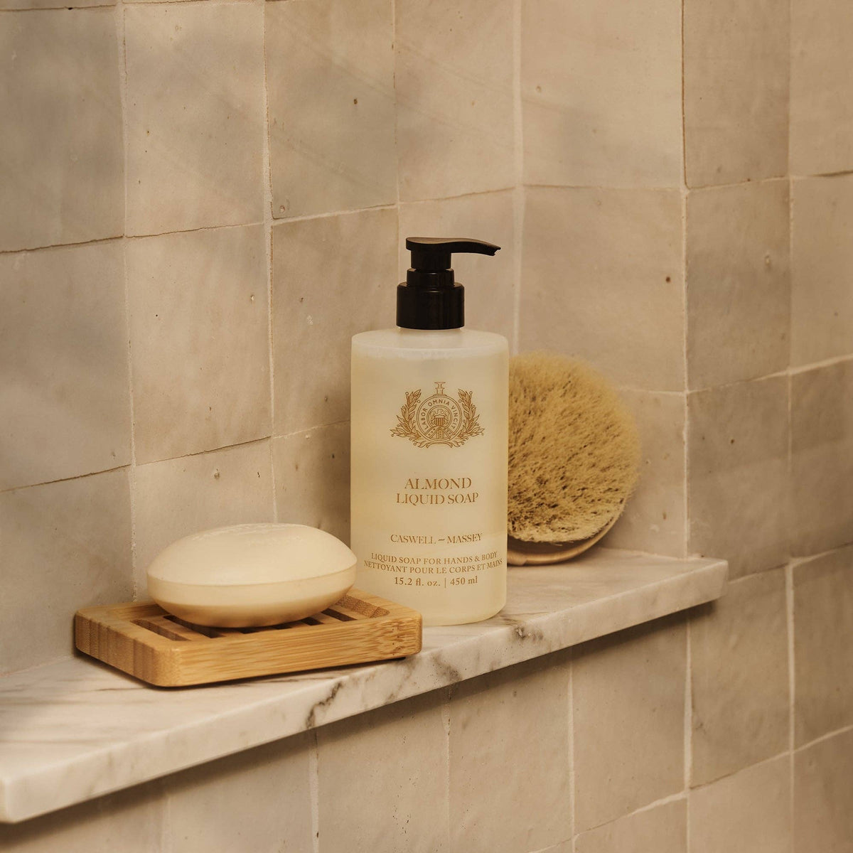 A marble shower shelf holds a Caswell-Massey Almond & Aloe Hand & Body Wash with pump, a round bar of soap on a wooden tray, and a natural bath sponge, set against beige tiled walls.