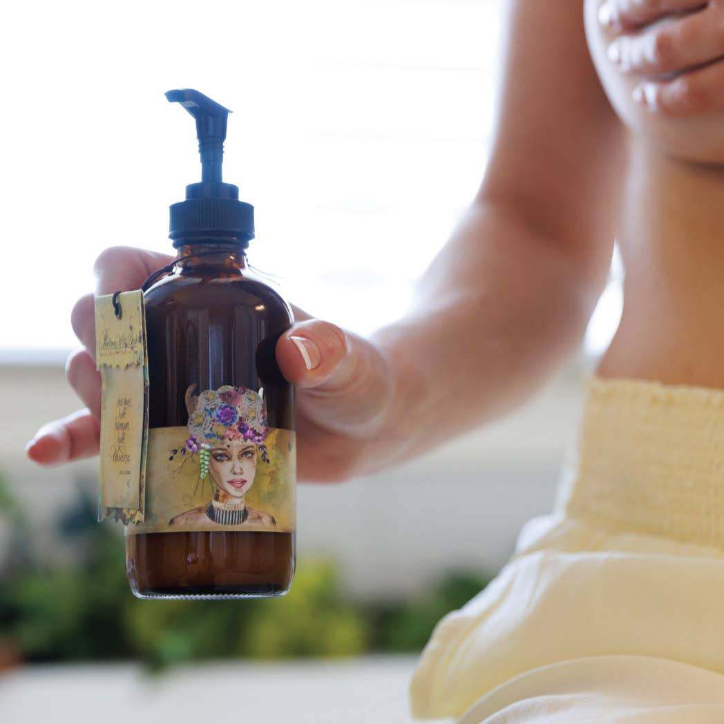A person holds a brown glass pump bottle of Barefoot Venus Lemon Freckle Body Cream, featuring a floral label with an illustrated woman in a flower crown; the softly blurred background adds emphasis.