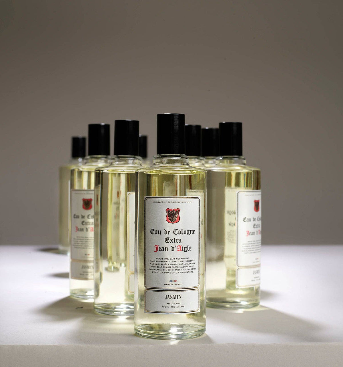 A collection of Jean d'Aigle Jasmine Eau de Cologne bottles are arranged on a white surface against a plain gray background. The clear glass bottles with black caps showcase a light yellow liquid with natural extracts and a white label featuring red and black detailing.