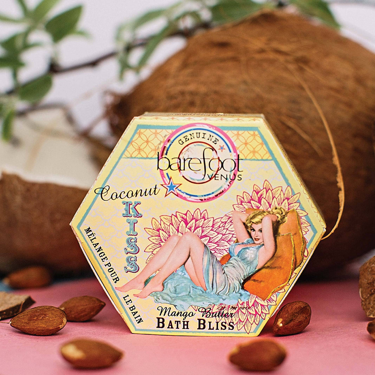 This image showcases a hexagonal box of "Barefoot Venus Coconut Kiss Bath Bliss Soak." The package features an illustrated woman lounging against a background of flowers. Surrounding the box are scattered almonds and a cracked coconut, emphasizing the natural ingredients used in this guilt-free indulgence.