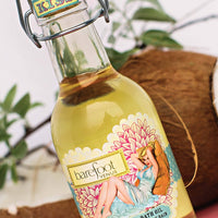 A bottle of Barefoot Venus Coconut Kiss Body + Bath + Massage Oil, by Barefoot Venus, is pictured against a backdrop of green foliage and a halved coconut. The bottle features a decorative label with an illustration of a relaxed woman surrounded by floral patterns, highlighting the blend of natural oils within.