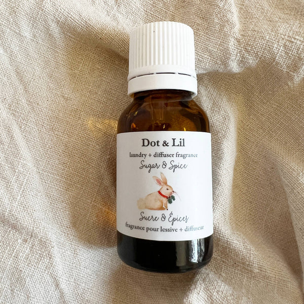 A small brown glass bottle labeled "Dot & Lil Sugar & Spice laundry + diffuser fragrance" features a bunny illustration and sits on textured beige fabric—a festive scent from Dot & Lil, evoking the cozy warmth of ginger cookies.