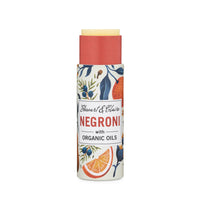A Stewart & Claire Negroni Lip Balm in a paper tube with organic oils, featuring a vibrant label with oranges and botanicals. The eco-friendly balm is partially twisted up, visible above the Stewart & Claire tube.