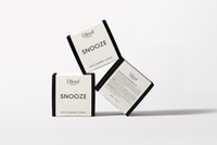 Three white and black L'floof SNOOZE Dog Calming Candle boxes are displayed: two show the product name, while the third highlights the essential oil formula on the back, designed to promote pet relaxation. Each candle contains 330ml.