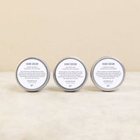 Three tins of Norfolk Natural Living hand creams labeled "hand cream" from the English countryside, each stating different essential oils and benefits (Coastal Walks, Rose Garden, En Plein Air), displayed side by side on a neutral background.