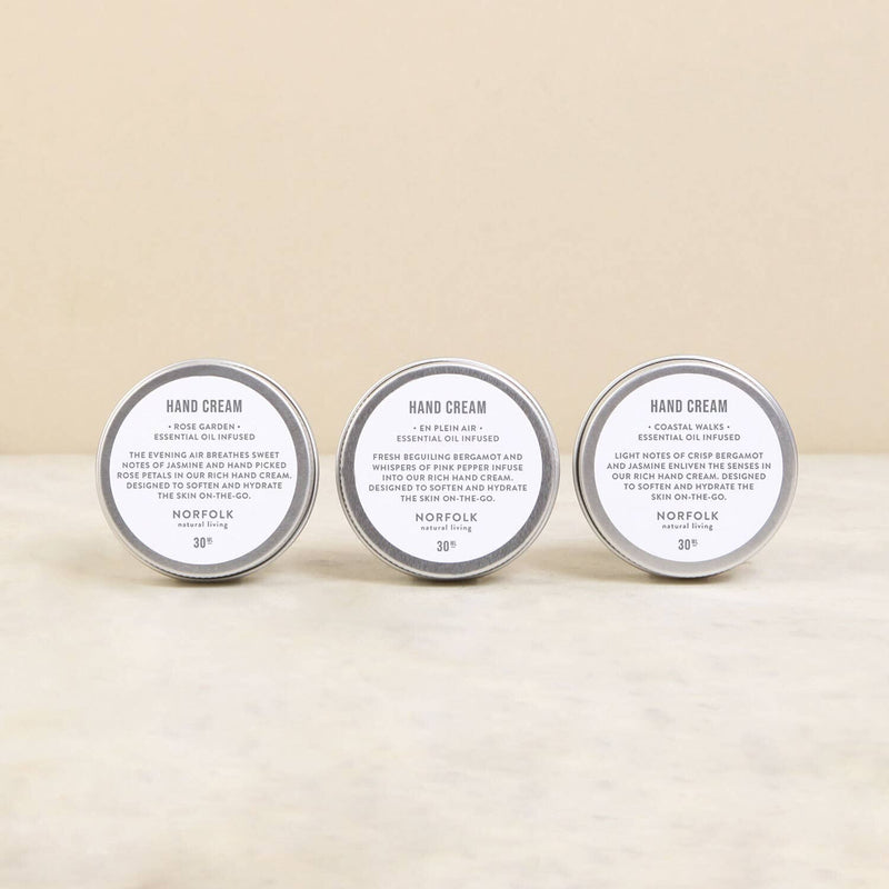 Three tins of Norfolk Natural Living hand creams labeled "hand cream" from the English countryside, each stating different essential oils and benefits (Coastal Walks, Rose Garden, En Plein Air), displayed side by side on a neutral background.