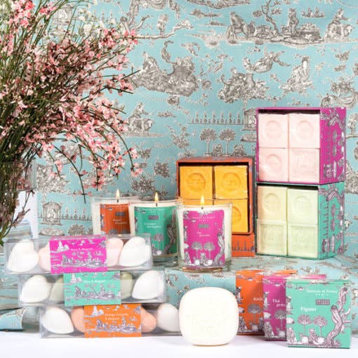 Senteurs De France Coffret Honeysuckle, Almond, Jasmine, and Verbena Soaps by Senteurs De France are displayed beside pink flowers, set against ornate blue and white wallpaper for a colorful luxury bath showcase.