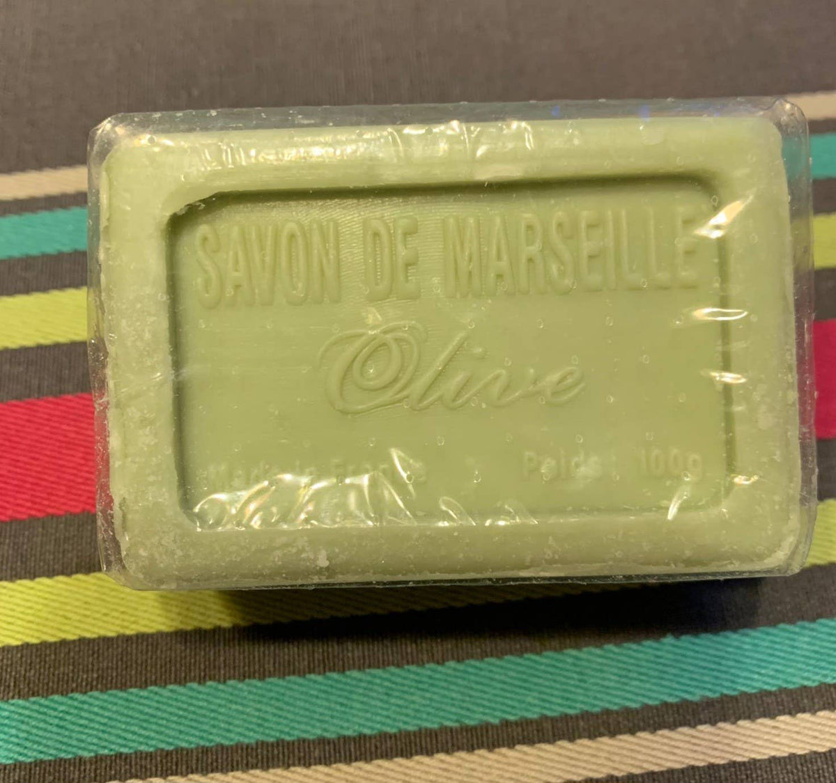 A green rectangular Senteurs De France Olive Oil Marseille Soap bar, with raised lettering and clear plastic wrap, sits on a colorful striped surface.