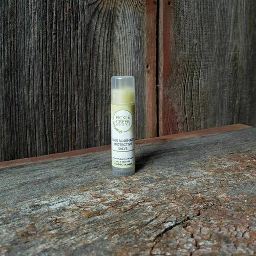 A tube of Pickle Creek Herbs Sage Rosemary Salve stands upright on a weathered wooden surface, with a rustic wooden wall in the background.