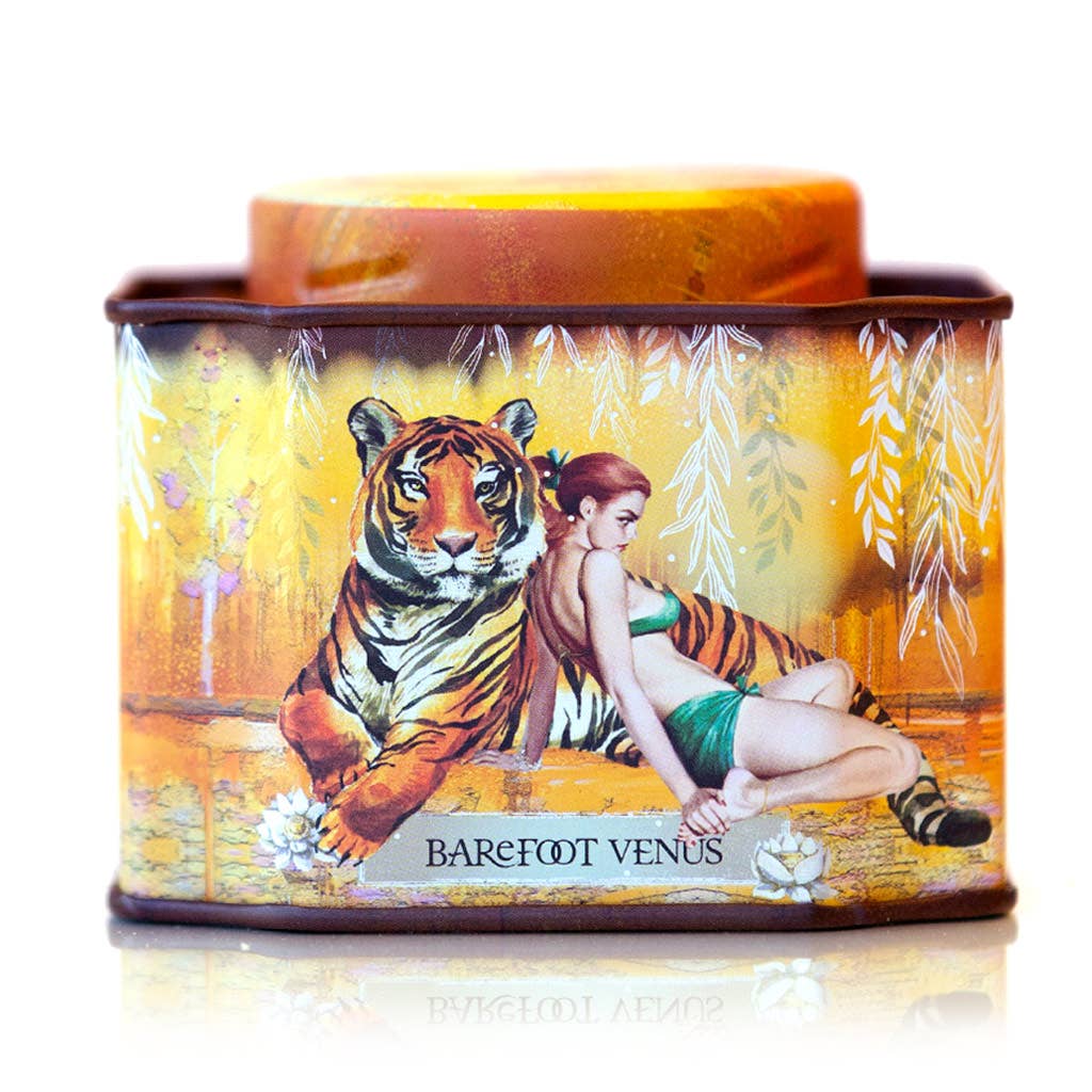 A decorative tin for Barefoot Venus Apricot Brandy Bath Soak features a woman in a green dress beside a tiger on a golden background with white leaves, combining handcrafted luxury with indulgent apricot scent.