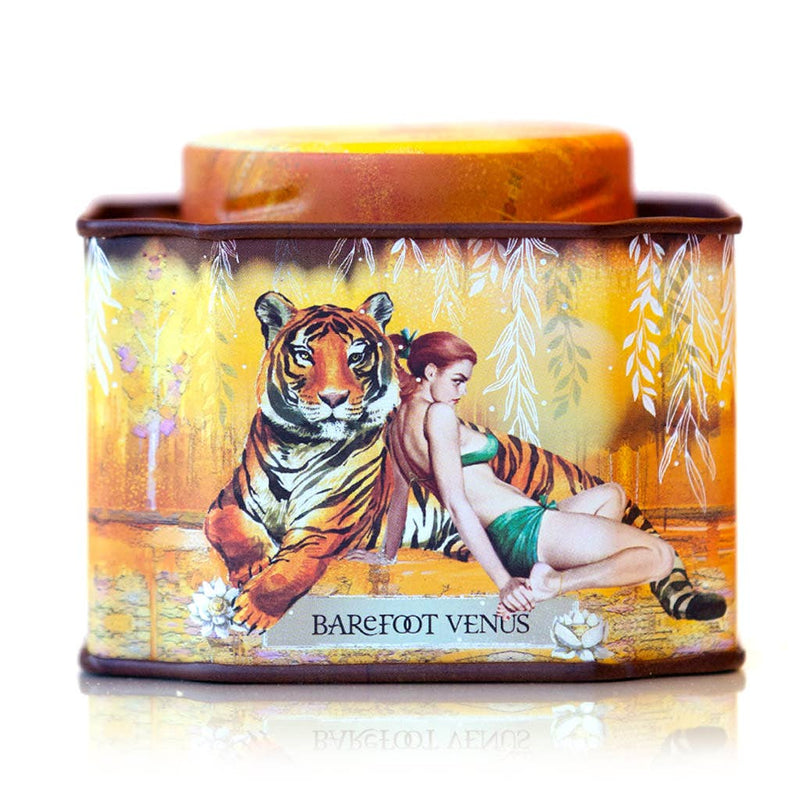 A decorative tin for Barefoot Venus Apricot Brandy Bath Soak features a woman in a green dress beside a tiger on a golden background with white leaves, combining handcrafted luxury with indulgent apricot scent.