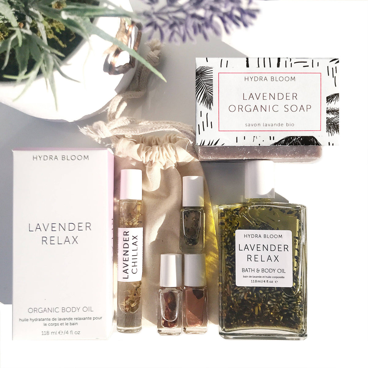 An assortment of Hydra Bloom Beauty Lavender-themed bath products, including organic soap, Hydra Bloom Beauty Luxe Lavender Bath and Body Oil, and small perfume rollers, displayed elegantly with natural light and a sprig of lavender.