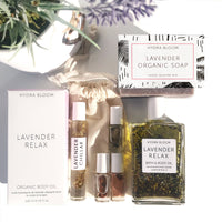 An assortment of Hydra Bloom Beauty Lavender-themed bath products, including organic soap, Hydra Bloom Beauty Luxe Lavender Bath and Body Oil, and small perfume rollers, displayed elegantly with natural light and a sprig of lavender.