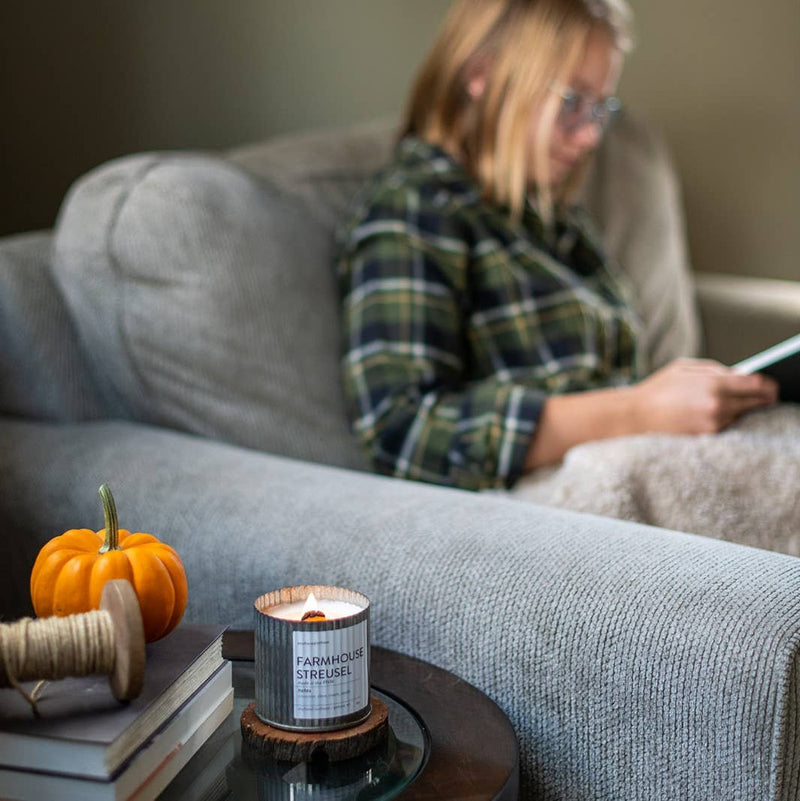 A person wearing glasses and a plaid shirt is reading a book while sitting on a gray couch. In the foreground, an Anchored Northwest Apples & Maple Bourbon Rustic Vintage Farmhouse Wood Wick Candle is lit on a table, accompanied by a small pumpkin and a spool of twine.