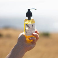A hand holding a transparent bottle of Nustad Family Ranch 4oz French Lavender Body Oil with a pump dispenser, against a blurry natural background.