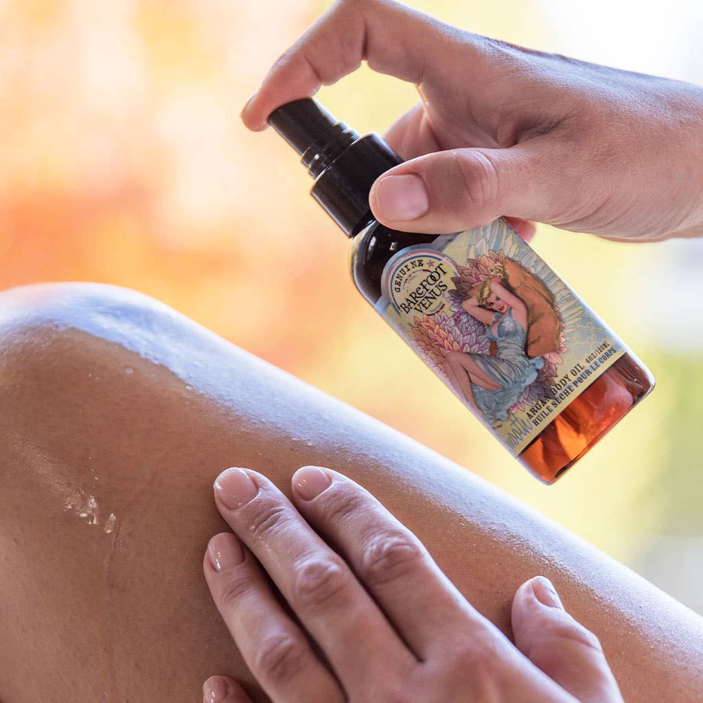 A hand is shown spraying Barefoot Venus Coconut Kiss Argan Body Oil by Barefoot Venus onto a person's leg, while the other hand gently spreads the liquid. The vividly labeled bottle, featuring a mythical character, is detailed in the softly blurred background of warm colors.