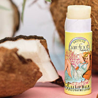 Close-up of Barefoot Venus Coconut Kiss Lip Balm placed beside a halved coconut, with a blurred background. The cruelty-free lip balm tube features vintage-style art of a woman in a blue dress reclining on a flower, evoking a tropical and nostalgic feel.