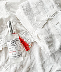 A bottle of Z&Co. Salt Farmhouse Room/Linen Spray with a red tassel sits next to a ribbon-tied white towel on a white fabric background. Handcrafted in Texas by Z&Co. for a fresh, inviting touch.