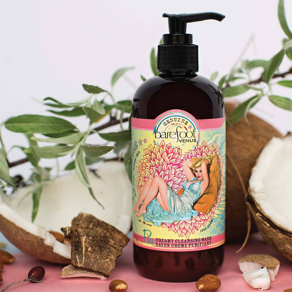 A bottle of Barefoot Venus Coconut Kiss Cleansing Hand & Body Wash is displayed against a backdrop of cracked coconuts and green leaves. The label features an illustrated woman lounging on a pink and yellow floral background. The bottle has a black pump dispenser, boasting an all-natural plant-derived formula.