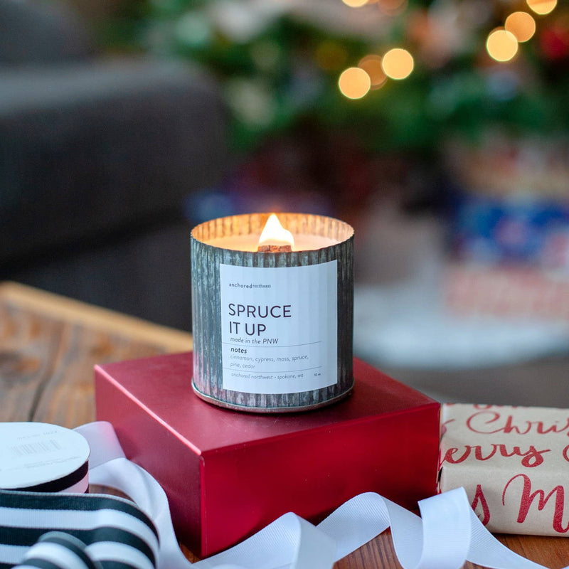 An Anchored Northwest "Mistletoe Kisses" rustic vintage farmhouse wood wick candle rests on a red box, with blurred holiday decorations in the background. Wrapped gifts and ribbons complete the festive scene.