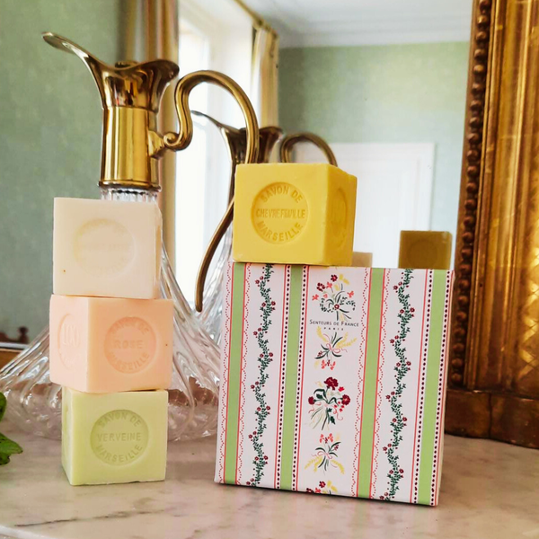 Three pastel Senteurs De France soaps—Rose Lily of the Valley, Verbena, and Honeysuckle—are stacked beside their floral box on marble, with a brass and glass pitcher plus mirror and curtains in the background.
