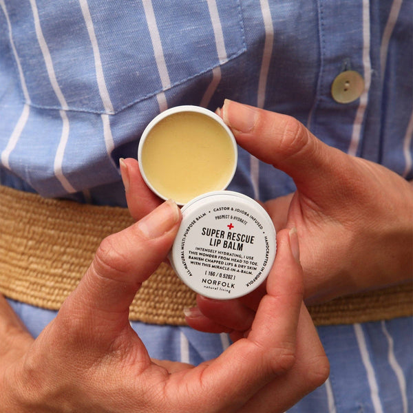 A person in a blue and white striped shirt holds an open tin of Norfolk Natural Living Rescue Lip Balm, showing the plant-based balm and the Norfolk Natural Living label on the lid.
