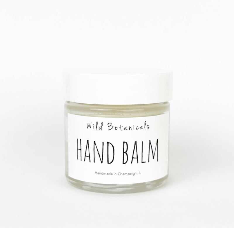 A small, clear glass jar with a white lid is labeled "Wild Botanicals Hand Balm 1oz Glass Jar". The text "Handmade in Champaign, IL" appears below the product name. The jar contains a cream-colored vegan hand balm made with organic Shea butter that promotes cell regeneration. The background is plain white.