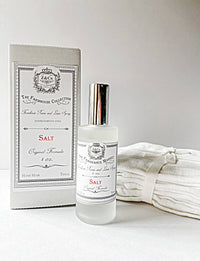 A clear glass Z&Co. Salt Farmhouse Room/Linen Spray stands before its matching white box, handcrafted in Texas and displayed with folded linens on a neutral background.