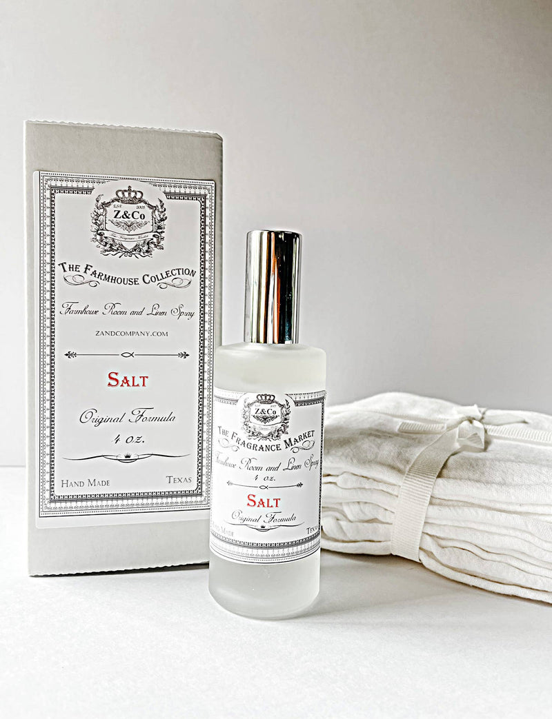A clear glass Z&Co. Salt Farmhouse Room/Linen Spray stands before its matching white box, handcrafted in Texas and displayed with folded linens on a neutral background.