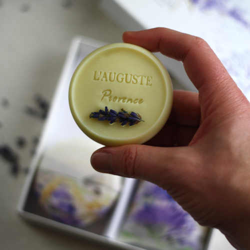 A hand holds a round, light yellow soap embossed with "L'AUGUSTE Provence" and topped with lavender. The background features an artistic gift box filled with four artisanal L'AUGUSTE Provence lavender soaps, adorned in a floral design reminiscent of lavender fields.