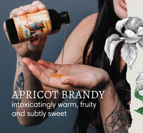 A tattooed person pours Barefoot Venus Apricot Brandy Bath Soak onto their hand. The text reads: "APRICOT BRANDY—intoxicatingly warm, fruity, and subtly sweet." Decorative silver leaves appear on the right.