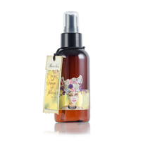 A brown amber spray bottle with a black cap and an attached tag showcasing artwork of a woman adorned with flowers and the words "Beneath His." Infused with the intense hydration of plant-based argan oil, its design boasts a mystical, artistic theme. The text on the bottle is not clearly readable. This product is known as Barefoot Venus Lemon Freckle Argan Body Oil from the Barefoot Venus brand.