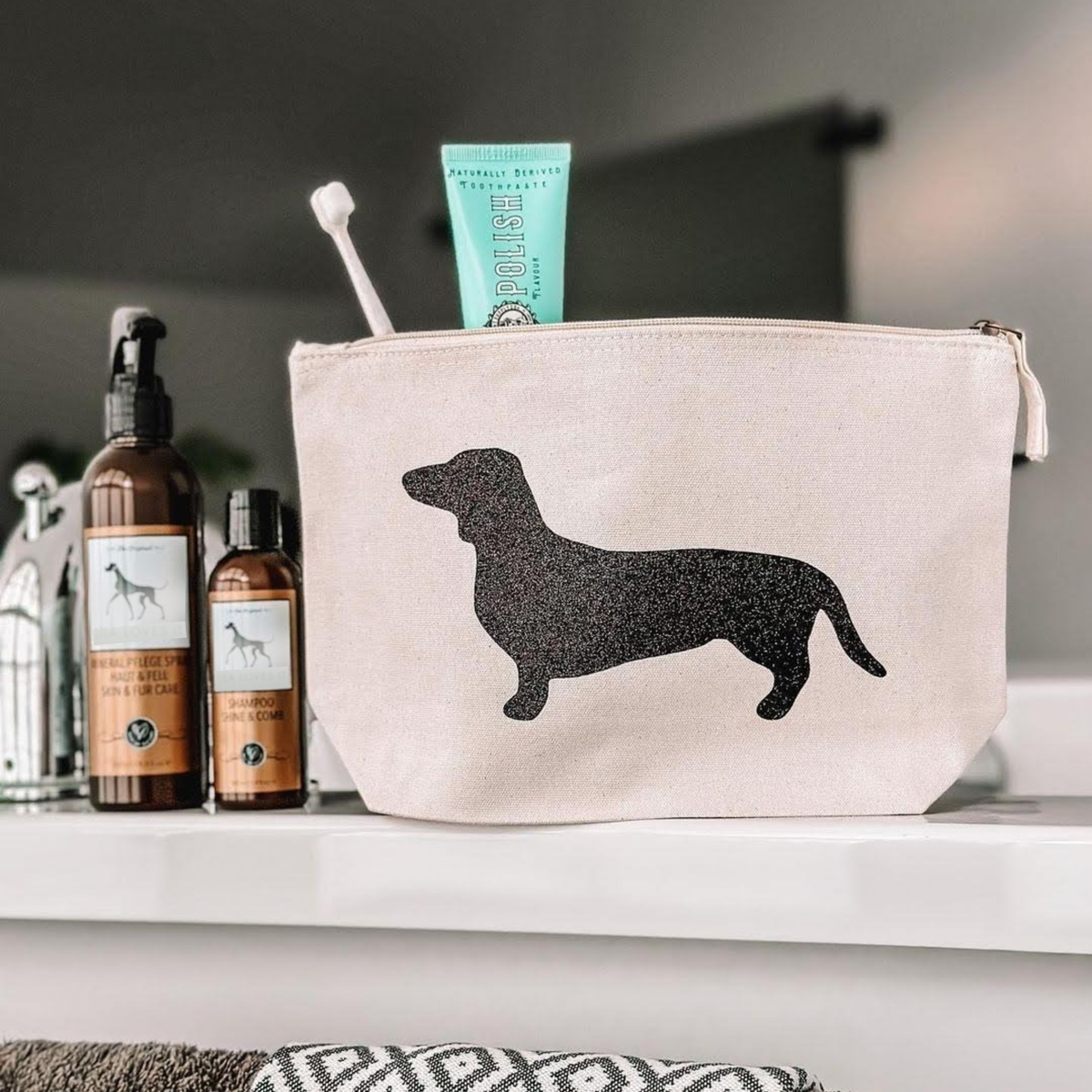 On a shelf sits a beige toiletry bag adorned with a black silhouette of a dachshund, containing the Wag & Bright Supply Co. - Puppy Polisher Eco Toothbrush (Medium), crafted from wheat straw bioplastic, and a tube of toothpaste. Beside it are two bottles of grooming products, each showcasing an illustration of a dog.