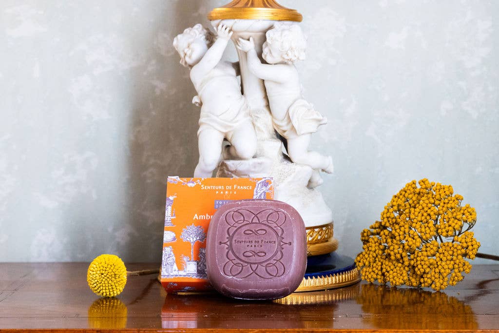 The Senteurs De France Ambre scented Soap - Toiles de Jouy, in its orange box, rests on polished wood with yellow flowers and cherub statues behind it, evoking French luxury.