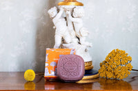 The Senteurs De France Ambre scented Soap - Toiles de Jouy, in its orange box, rests on polished wood with yellow flowers and cherub statues behind it, evoking French luxury.