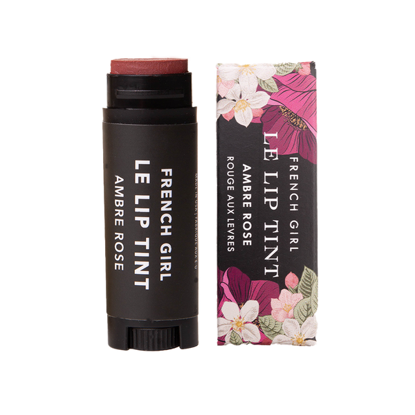 A tube of FRENCH GIRL Tinted Lip Balm in Ambre Rose stands beside its floral box, both showing the brand and product name in white on black—an organic balm giving lips a hint of color and hydration.