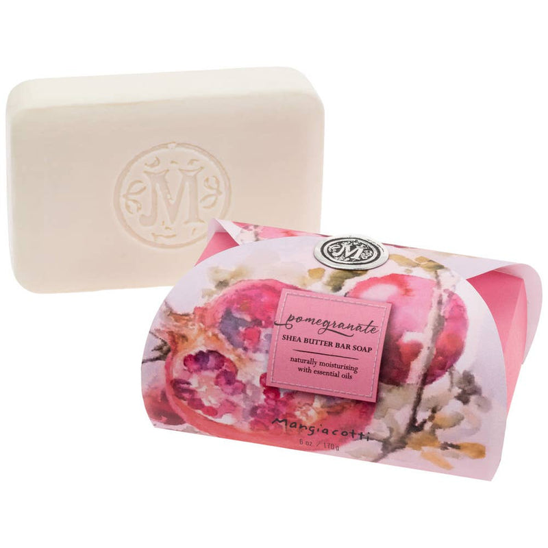 A **bar of Mangiacotti Pomegranate Shea Butter Bar Guest Soap 6 oz** with an embossed "M" on it is displayed alongside its packaging. The pale pink wrapper, adorned with a pomegranate design, features a label that reads, "**Mangiacotti Pomegranate Shea Butter Bar Guest Soap 6 oz**," highlighting its naturally moisturizing properties and essential oils.